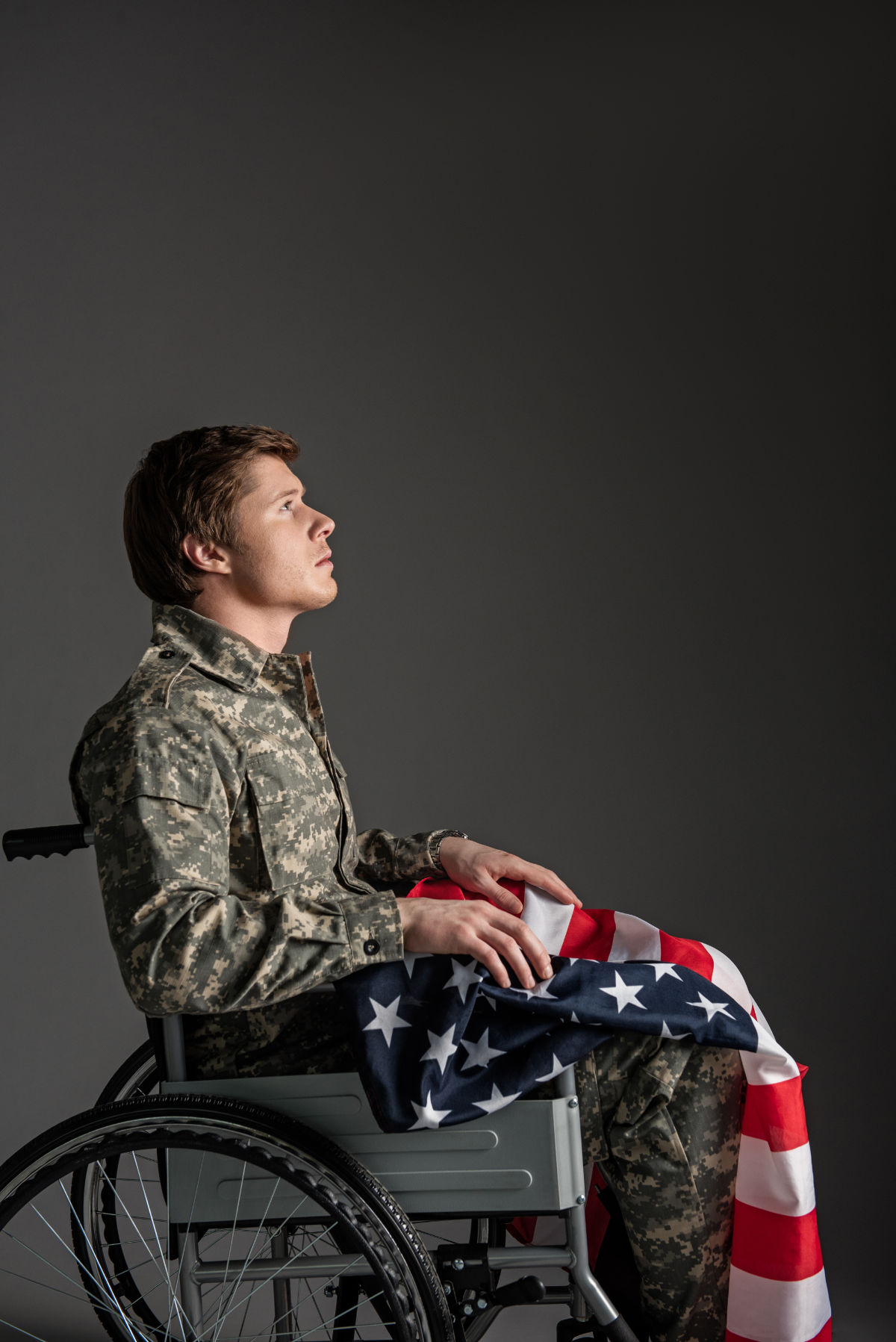 Sorrowful,Paralyzed,Soldier,Sitting,In,Wheelchair,With,Sad,Look.,He Sorrowful paralyzed soldier sitting in wheelchair with sad look. He is looking up and having a flag on his knees. Isolated on grey background